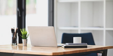 Photo of notebook, diary, coffee cup, pencil in vase, potted plant and computer laptop putting together on wooden working desk over modern living room windows and bookshelf as background.