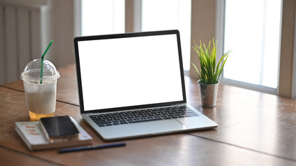 Photo of computer laptop with white blank screen putting on wooden table with take away plastic cup, notebook and smartphone over orderly living room as background.