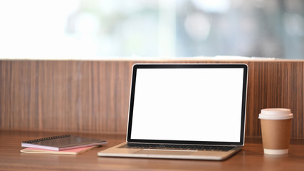 Photo of computer laptop with white blank screen putting on wooden table with coffee cup and notebook over orderly living room as background.