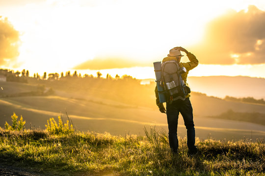Young man trekker in tuscany hills at sunset. Yellow jacket, backpack, hat. Traveling Volterra, Italy.