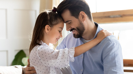 Head shot close up side view affectionate little child daughter touching foreheads with smiling bearded father. Happy small kid girl enjoying sweet moment with dad in living room, trustful relations.