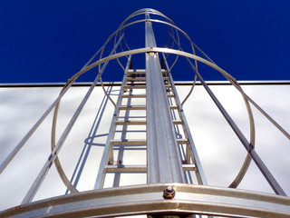 aluminum roof access ladder with safety cage in diminishing perspective. strong shadows on white concrete wall. rungs and vertical safety bars. buildings, architecture and technology. clear blue sky. © Istvan
