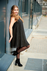 Photo of a fashionable blonde girl in a dress on a background of a wall of an outdoor building in a city in spring