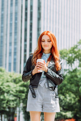 Happy young trendy red hair woman drinking take away coffee and walking in an urban city.
