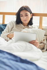 Vertical portrait of modern adult woman using digital tablet and browsing internet while lying on comfortable bed in morning, copy space