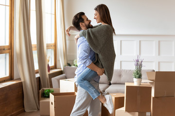 Strong affectionate young man in glasses lifting smiling wife in living room in new apartment...