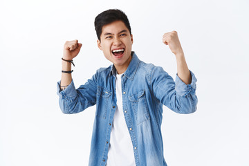 Portrait of empowered, enthusiastic asian man fist pump, raise hands up in triumph, celebrating victory, smiling saying yes, winning game or competition, achieve goal, white background