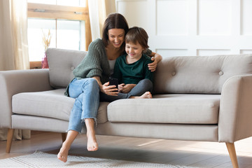 Fototapeta premium Full length smiling young barefoot woman hugging small cute child, taking selfie photo together on smartphone. Pleasant happy mommy resting on sofa with little kid boy, showing funny mobile apps.