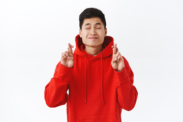 Portrait of hopeful, dreamy young asian man have wish, cross fingers good luck, close eyes and pleading with delighted, longing face, dreaming about something badly, white background