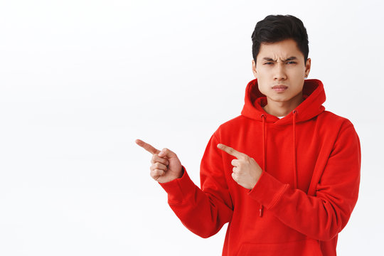 Portrait of young asian man looking with judgement and disappointment at camera, frowning angry pointing fingers left, demand answers, being displeased and outraged with something, white background