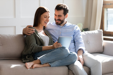 Excited young married couple resting on sofa, watching comedian funny movie on computer tablet, enjoying free weekend leisure time together. Happy family spouses cuddling, shopping online at home.