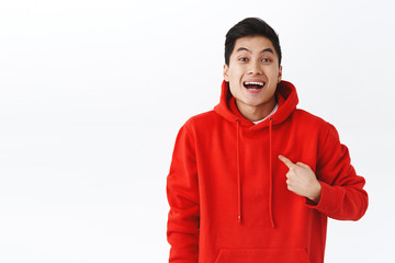 Portrait of excited, happy smiling man in red hoodie pointing at himself as being chosen or picked, achieve goal, talking personal goal, standing cheerful white background