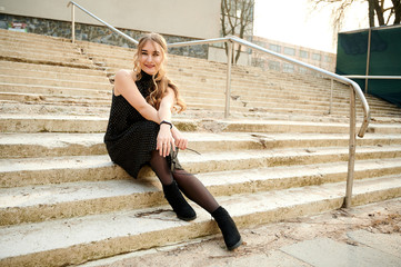 Photo of a fashionable blonde girl in a black dress sitting in various poses on the steps of an outdoor building in the city in spring