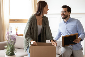 Overjoyed young married couple unpacking belongings from cardboard boxes, moving into new apartment flat. Happy 30s woman unboxing things, decoration items with smiling bearded husband in glasses.