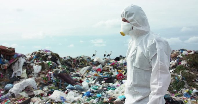 Virologist in protective costume and gas mask walks near landfill dump site
