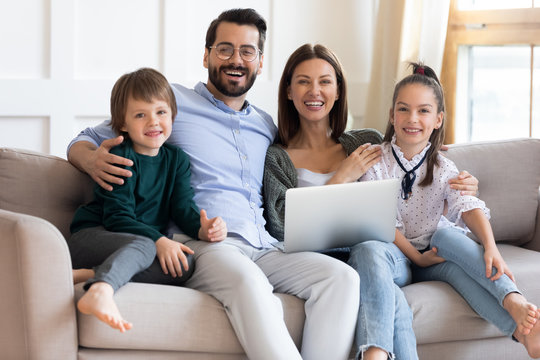 Portrait of happy full family with kids relaxing on couch with computer. Smiling married couple embracing small children, posing for photo in living room, looking at camera. weekend pastime.