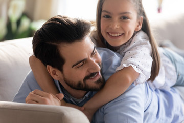 Head shot close up handsome young father holding little schoolgirl on back, enjoying sweet moment in living room. Smiling small kid girl lying with happy dad on sofa, warm relations concept.