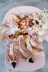 a wedding bouquet of flowers and greenery stands on a pink chair next to model women's shoes