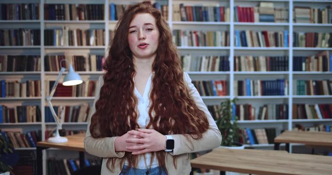 Portrait of beautiful young red head girl emotionally speaking and making a lot of gests by her hands in library. Bookcase in background.