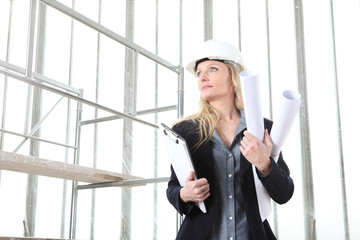 woman architect or construction engineer wear helmet and holds folder and blueprint inside a building site with windows and scaffolding in the background