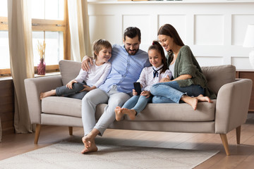 Full length happy young family couple relaxing on comfortable sofa with smiling two little children siblings, using smartphone. Joyful parents spending free weekend time with kids in living room.