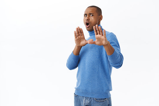 Portrait of young reluctant nervous african-american man telling stay away from me, making block with raised hands, stop gesture, step back from cringy person, refusing, white background