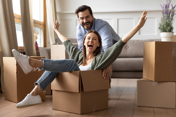 Full length overjoyed young bearded man in glasses pushing laughing wife in carton box. Energetic...
