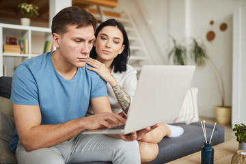 Fototapeta premium Portrait of modern young couple using laptop together while sitting on sofa in cozy home interior, online service concept, copy space