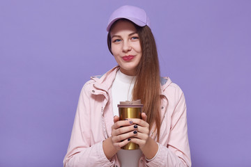 Close up portrait of charismatic charming attractive young female holding themo mug in both hands, drinking coffee, having walk, wearing pink cap and leather jacket, white sweater. Youth concept.