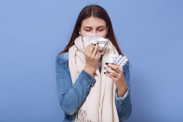 Picture of ill isolated brunette blowing her nose, holding paper napkin, using nasal drops, closing eyes, wearing jeans jacket and scarf, taking pills, being on sick leave. Health problems concept.
