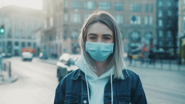 Young serously woman in medical mask standing in city street looking to camera road with cars on background Concept of health and safety life COVID-19 coronavirus virus protection pandemic in world