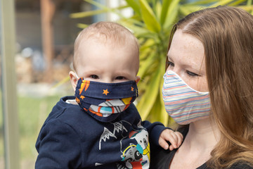 Portrait of baby and mother together with reusable protective homemade face mask. Baby boy is looking at the camera and mother is looking at her baby. 