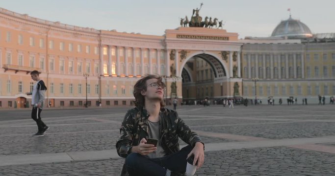 The beautiful young man siting on the Palace square and drinks coffee at sunset, he dressed in a military jacket and jeans, long curly hairs, Arch of the General Staff Building is on background