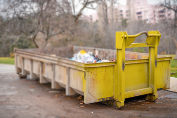 Yellow dumpster seen on a street near a park. The dumpster is filled to the top with plastic trash bags and other debris. Selective focus.