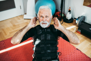 Good looking and positive senior man doing exercises in electrical muscular stimulation suit. - obrazy, fototapety, plakaty