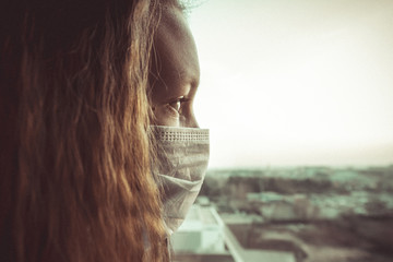Girl wearing face mask because of virus epidemic in the city. Young woman looks window in medical mask, protect from infection of coronavirus, pandemic, outbreak of disease. Global quarantine concept