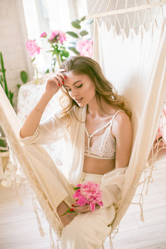 Cute tender young girl with blond long hair surrounded by pink peonies is resting in a hammock in the studio. Spring and flowers. Beauty and Fashion