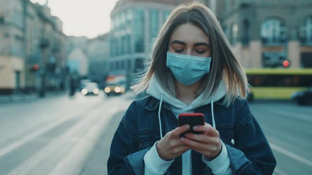 Young blonde woman in protective medical mask walks down to the street uses phone texts scrolls surfs the internet search news covid19 coronavirus virus protection pandemic city slow motion close up