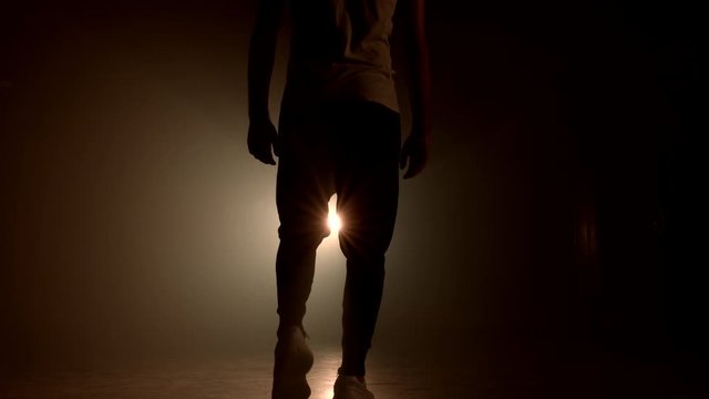 Silhouette of young man in sports wear is walking through smoky studio or scene right from camera. Backlight, bright spotlight on background.