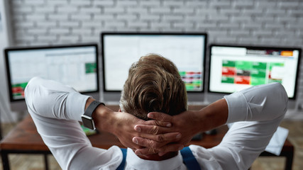 An awesome financial state of mind. Rear view of middle-aged trader sitting back in front of pc and analyzing stock market chart, while working in the office. - obrazy, fototapety, plakaty