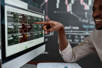 Utilising skills for earnings. African businessman, trader sitting in front of computer screen and looking at graph chart with a smile. Blackboard full of data analyses in the background.