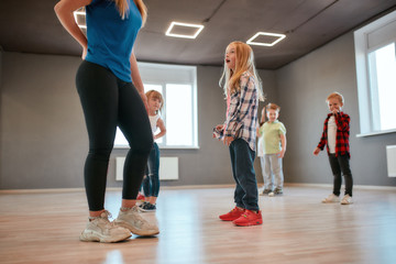 Fototapeta premium Cute little girl is talking with her female dance teacher while standing in the dance studio. Group of cute little boys and girls studying modern dance in choreography class