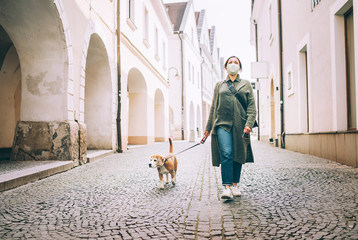 Young female fancy-dressed using a face mask as a coronavirus spreading prevention walking with her beagle dog on deserted old European squares and streets. Global COVID-19 pandemic concept image.