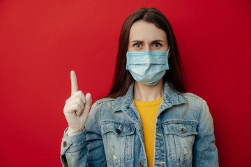 Young serious woman model wears protective medical mask, points away, demonstrates something at blank copy space for your advertisment or promotional text, looks at camera, isolated over on red wall.