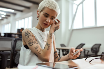 Enjoying free time. Young cute blonde tattooed business woman putting on wireless earphones while...