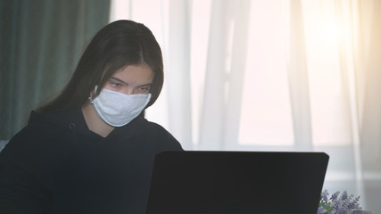 young girl in protective anti virus face mask works on laptop at home