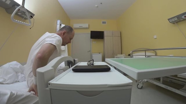 A man in white hospital pyjamas gets out of bed and sends a message and goes back to bed. On the bedside table are a smartphone and glasses. In the background is a switched off TV.