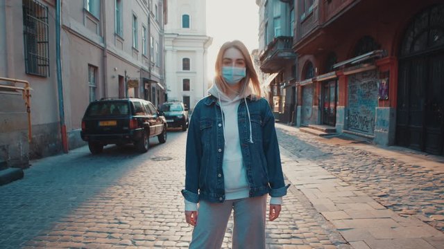 Young blonde european woman wearing protective medical mask looking to camera in old city street sunset sunlight to the background covid19 corona virus protection pandemic city slow motion outdoor
