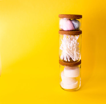 cotton swabs, colored cotton wool ball, cotton discs inside glass small container with wooden top. Yellow background. Free copy space.