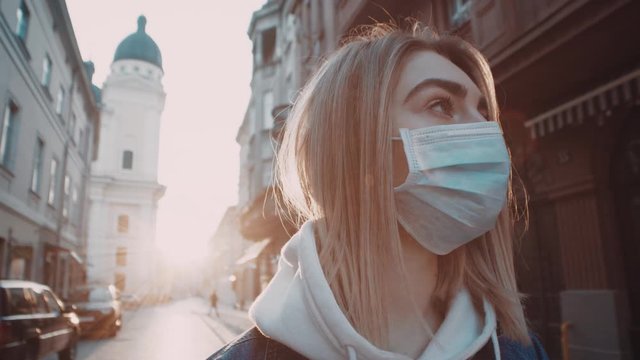 Extrime close up view of young blonde woman in protective mask stands in city street looks around sunset sunlight to the background covid19 corona virus protective pandemic city slow motion outdoor